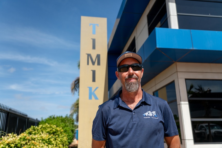 A man in a navy blue cap stands in front of a building with a logo that reads TIMIK. 