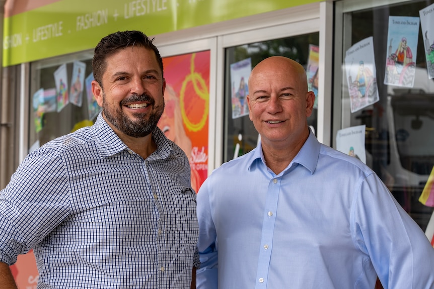 A man with a a beard and another man stand together looking at the camera.