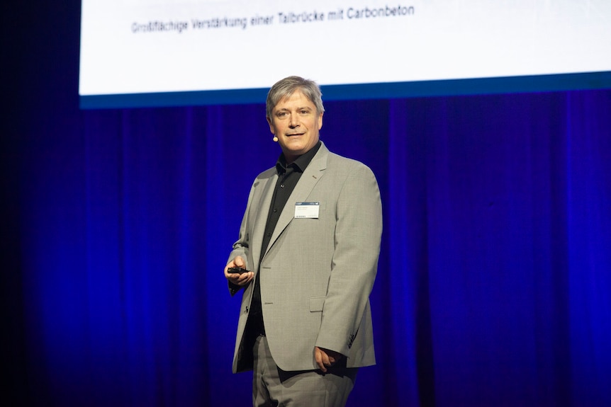 Colin Caprani stands on a stage in front of a blue curtain, wearing a beige suit.