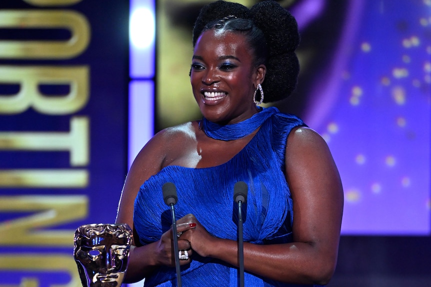 A smiling Wunmi Mosaku, dressed in a royal blue evening gown, speaks into a microphone on stage.