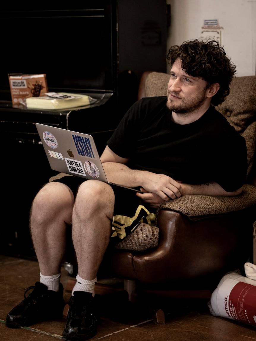 Declan Greene, early 40s, sits in a 70s-style chair in rehearsal room, laptop on his lap, leaning to the side.