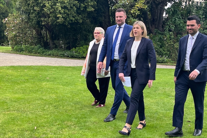 Two women and two men walk across green grass with trees behind them.