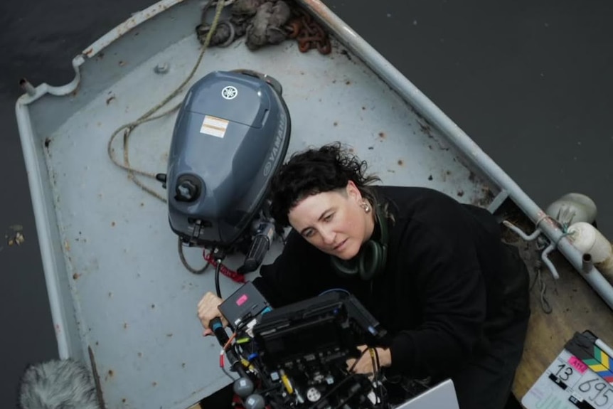 Woman wearing dark-coloured clothing, and has short dark hair, kneels down while looking into the back lens of a camera