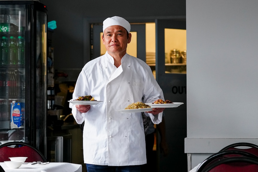A male chef walking while holding three plates of dishes on top of his arms