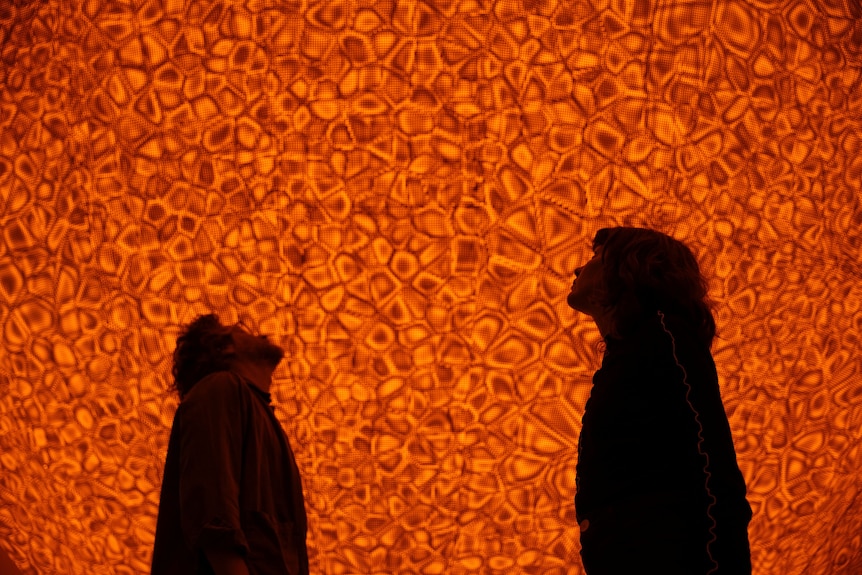 Two people look up at an orange installation of textured shapes, made from light, steel, aluminium, foil and glass.