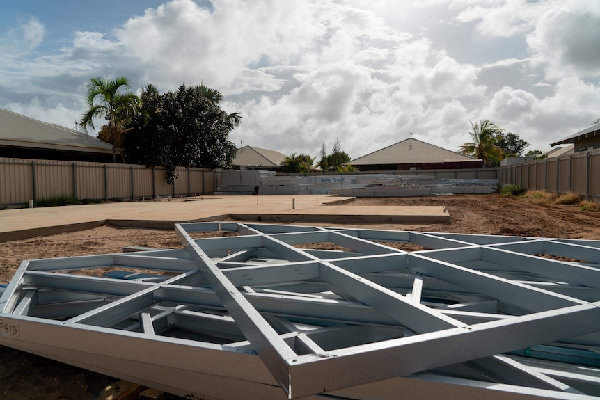 Steel frames lie on red earth on an empty block.