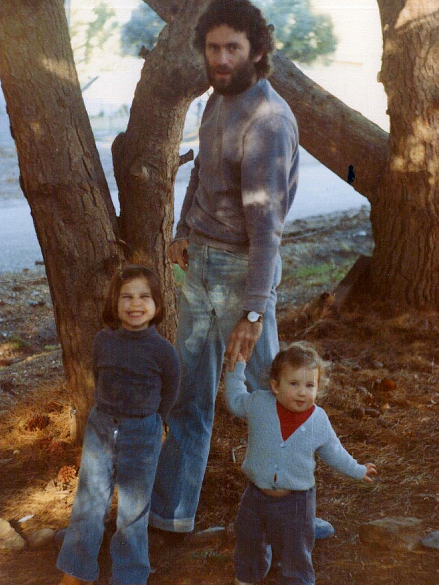 A father and his two toddler daughters stand outside under a tree dressed in 1970s attire. The younger girl is mid-run