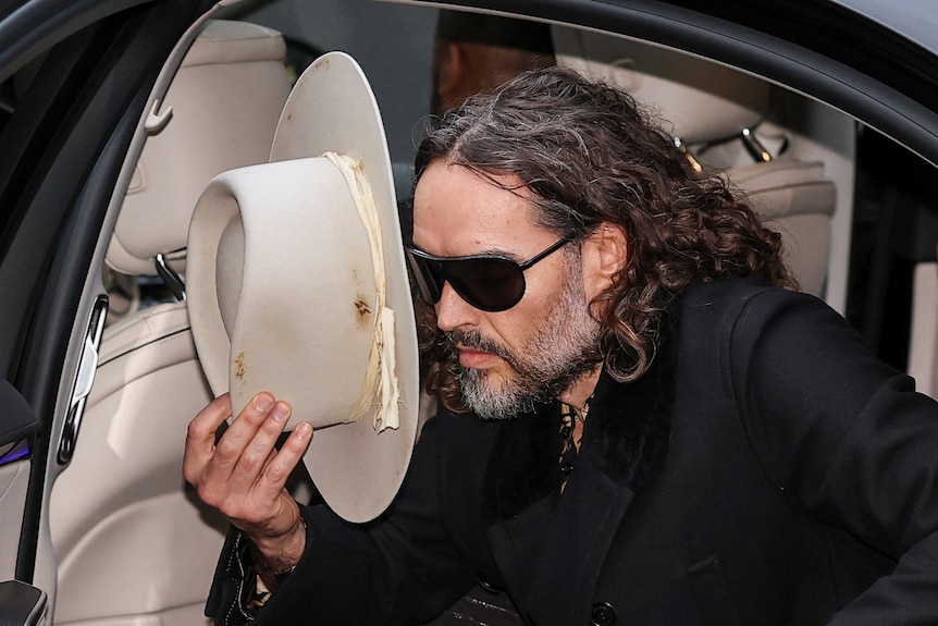 Russell Brand leans forqard to step out of a car, wearing sunglasses and holding a white cowboy hat to shield his face
