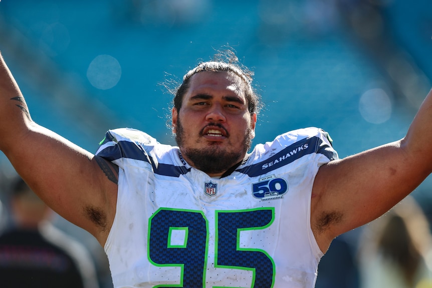 A Samoan man in a white Seattle Seahawks football jersey looks toward the camera with his hands raised.