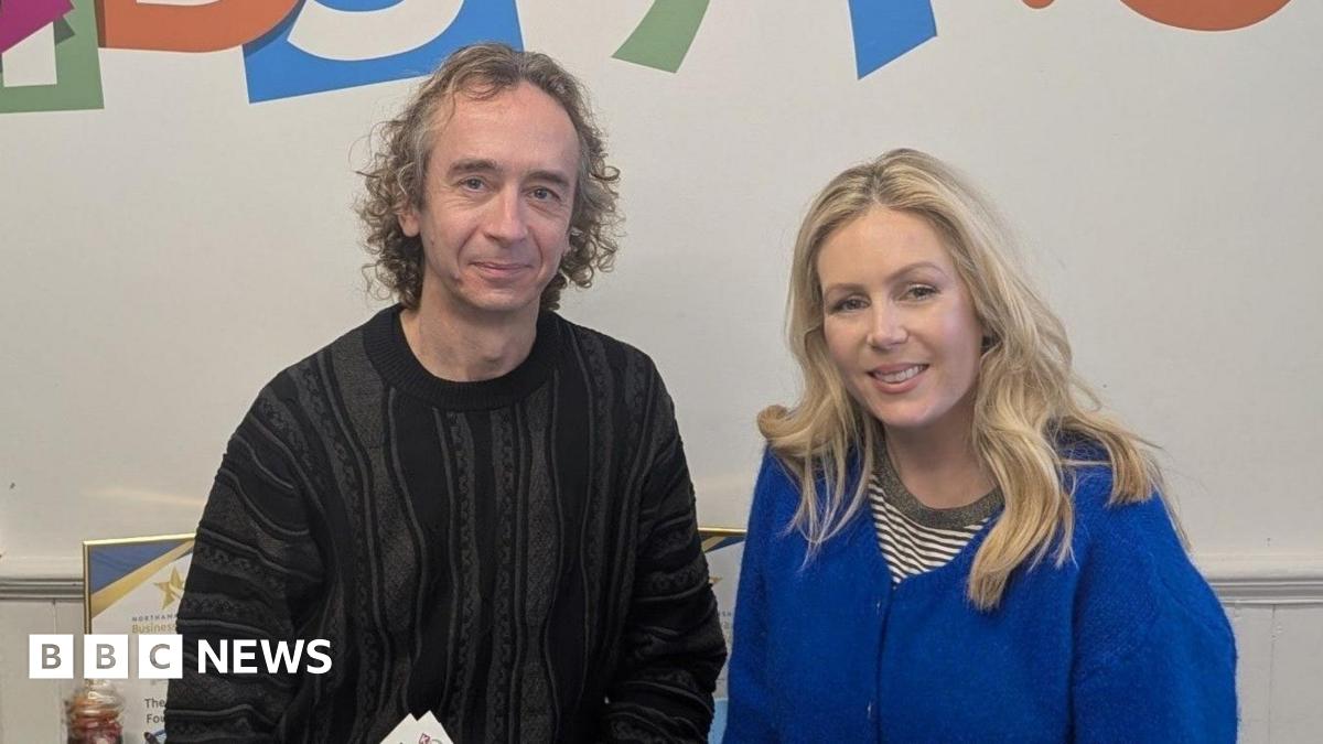 Geoff Russell-Jones in a black jumper with curly hair smiles at the camera while standing next to Rebecca Caswell-Fox, who has long blonde hair and a blue cardigan.