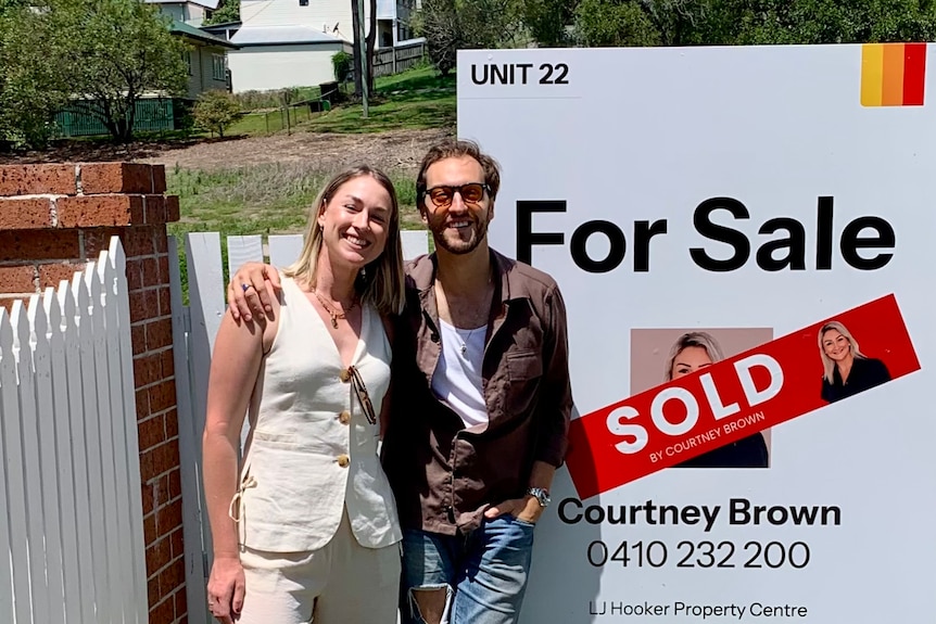 A man and woman in their early thirties stand smiling in front of a 'for sale' sign