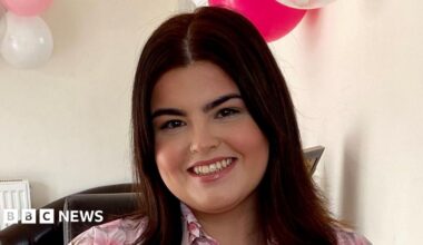 Ayla Jones is a white young woman with shoulder length dark brown hair. She is wearing a floral pink blouse. Behind her on the wall are some clear, white and pink balloons and there is pink confetti on the table. She is smiling directly at the camera.