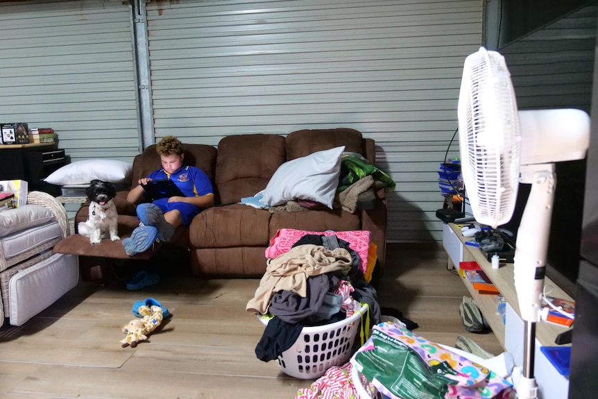 A boy in a school uniform sitting on a couch in a shed with a white fan angled towards him.