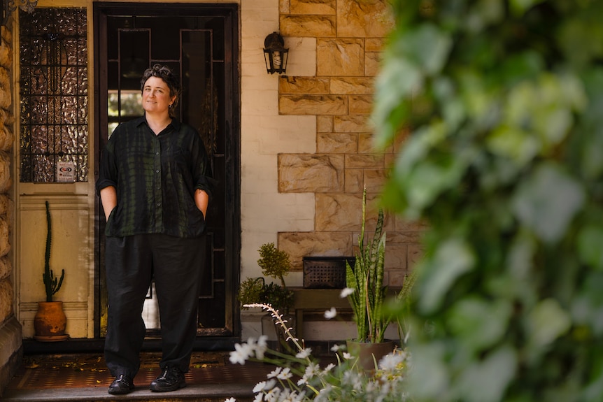 A woman with short dark hair wearing all black smiles slightly standing with hands in pockets at front door entrance of house
