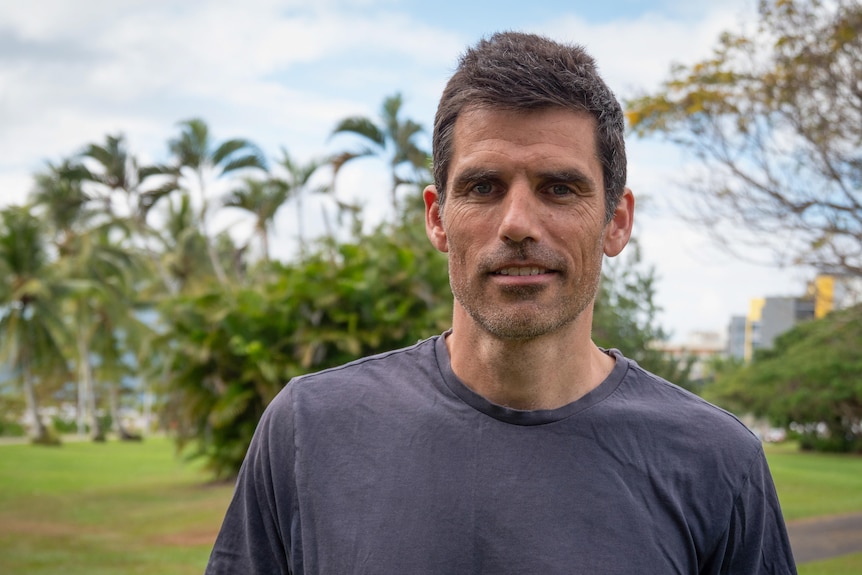 Outback Mike, wearing dark T-shirt, smiles to camera standing outside with palm trees behind