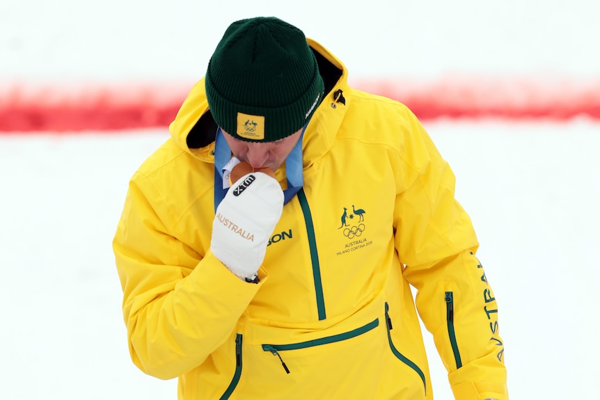 A skier in a yellow snow jacket and beanie kisses a gold medal