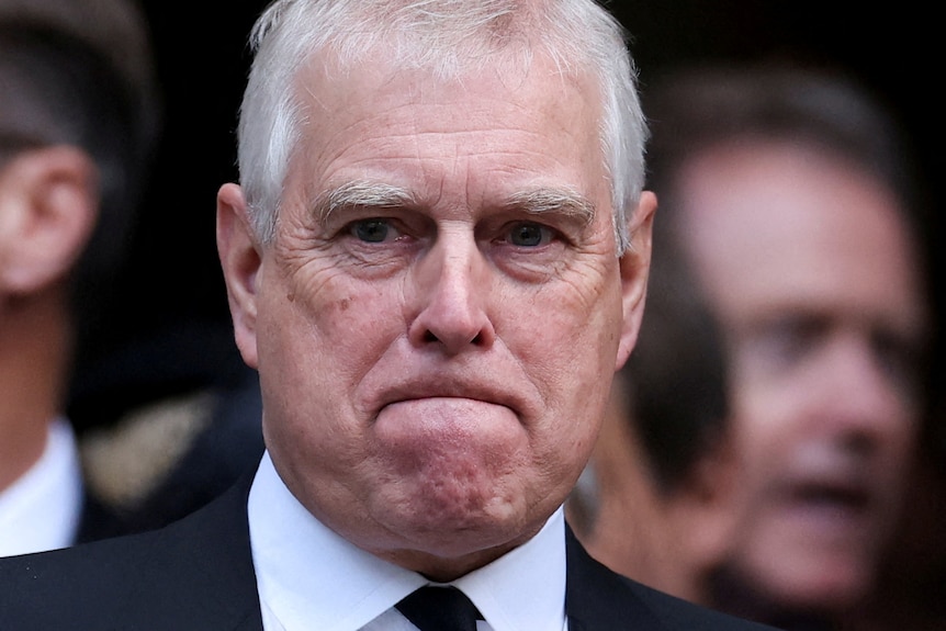 Prince Andrew looks unhappy as he is dressed in a dark suit surrounded by a crowd of people.