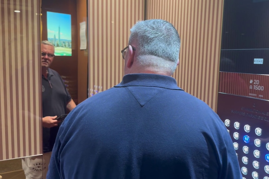 A man with a navy shirt stands in an elevator with his back to the camera. A mirror reflects his face showing he has glasses.