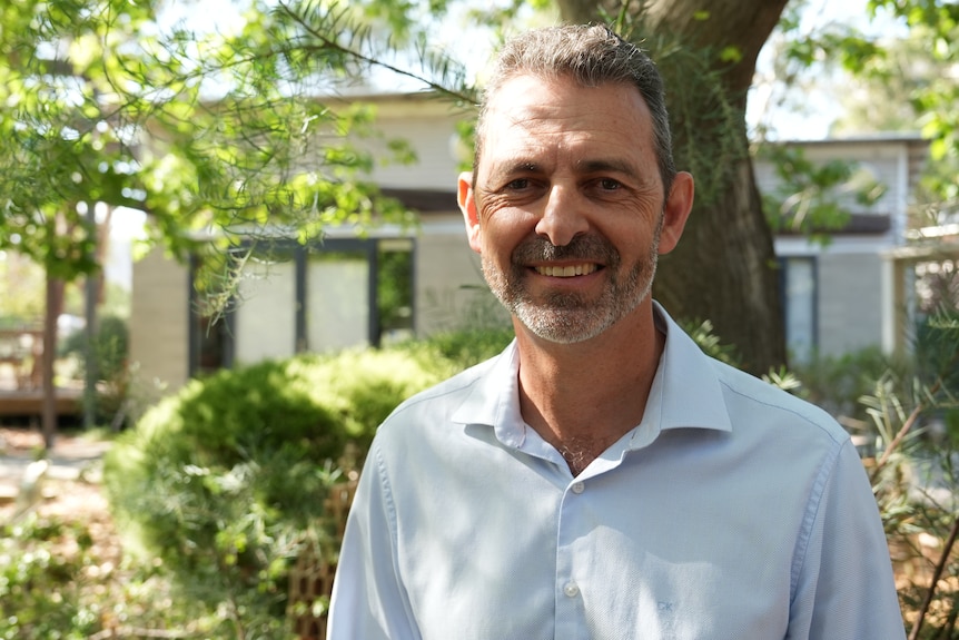 Mark stands outside among trees, wearing a collared shirt and smiling.