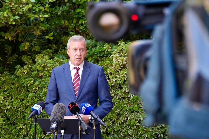 A mid-shot of WA Police Minister Reece Whitby speaking at a media conference outside, with a TV camera in the foreground.