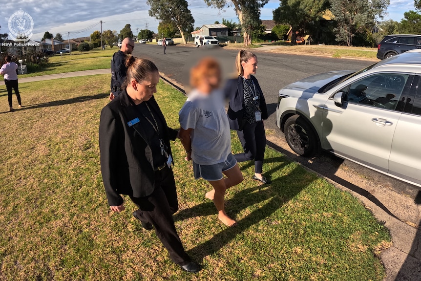 A woman in shorts and a T-shirt is escorted along a suburban street by plainclothes police.