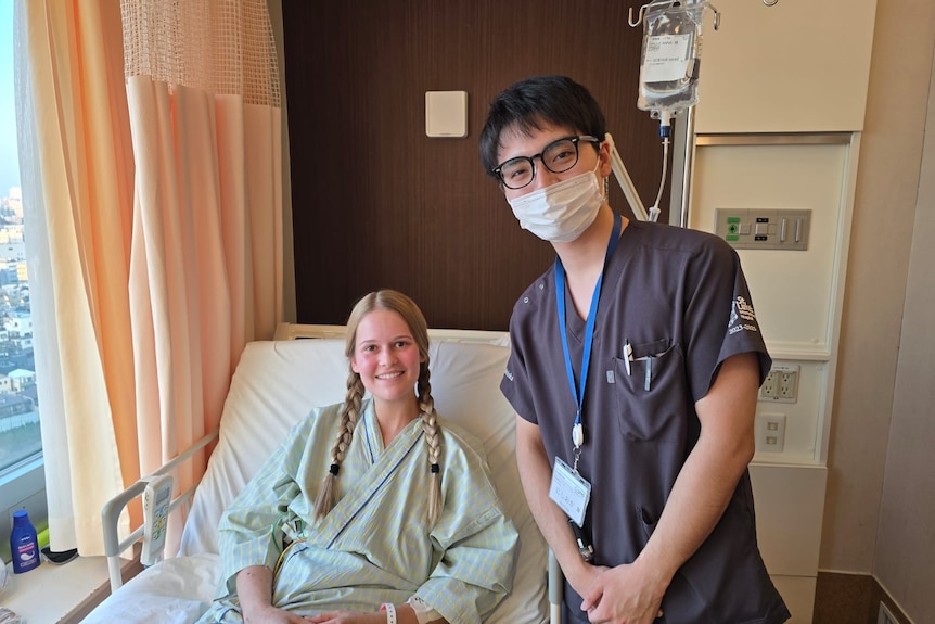 A young woman in hospital gown sits up in bed smiling next to a Japanese doctor in dark scrubs.