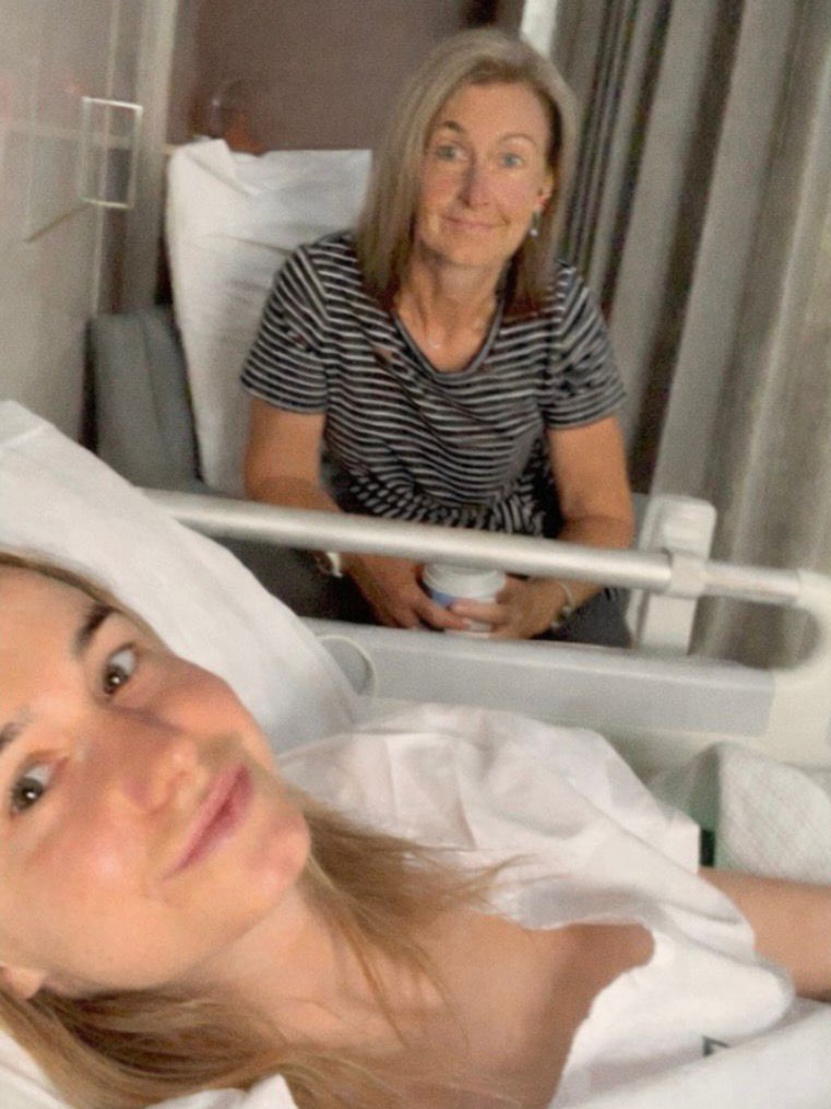 A young woman smiles in a selfie taken as she is lying in a hospital bed. A woman sits next to the bed also smiling.