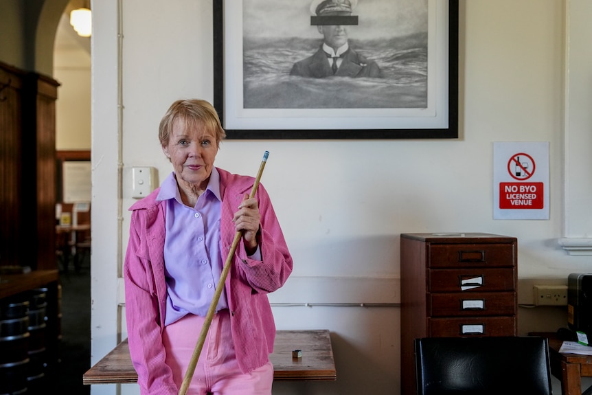 Volunteer Robbie Murphy wears a pink top and pants and holds a pool cue
