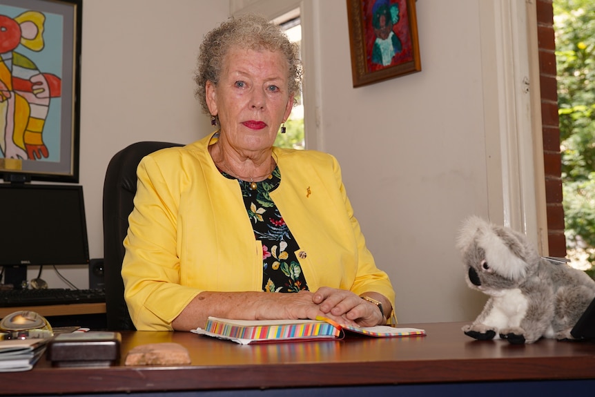 A senior lady with curly hair wearing a yellow blazer sits at a desk. There is a soft toy Koala on her left.