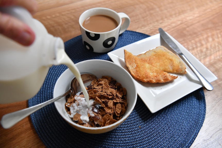 a breakfast spread on a dining table. a person is pouring milk into a bowl of cereal