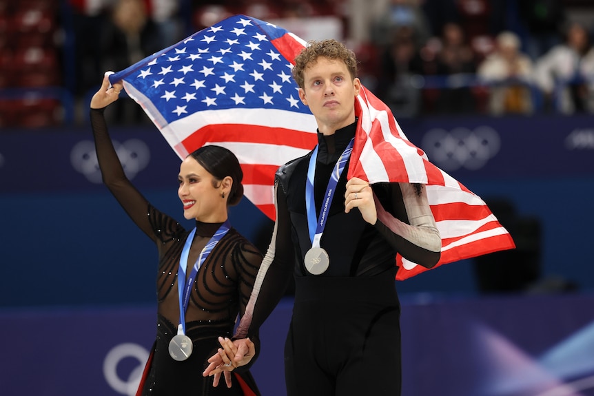 A man and a woman hold a US flag over there heads, walking with silver medals around their necks
