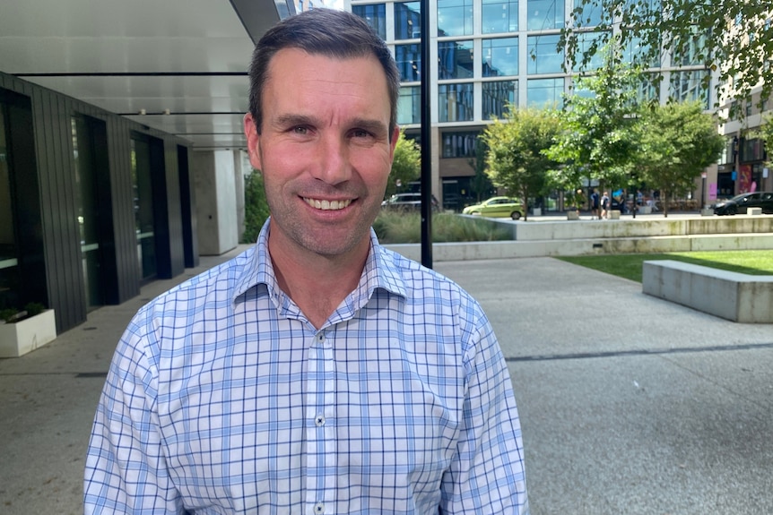 A man with short brown hair wearing a button down shirt stands outside an office building smiling.