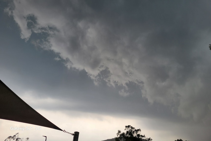 Dark grey clouds hang low with darker skies behind them above a edge of a dark shade sail and a tree.