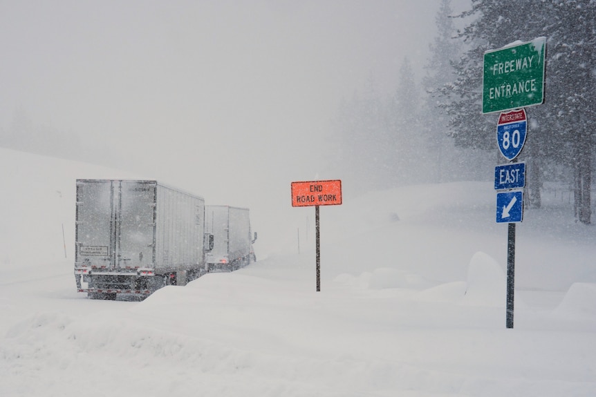 Two trucks covered in snow with an orange road sign and a green road sign next to them.