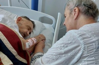 An older man lies in a hospital bed under a bright quilt, holding the hand of a woman sitting beside him.