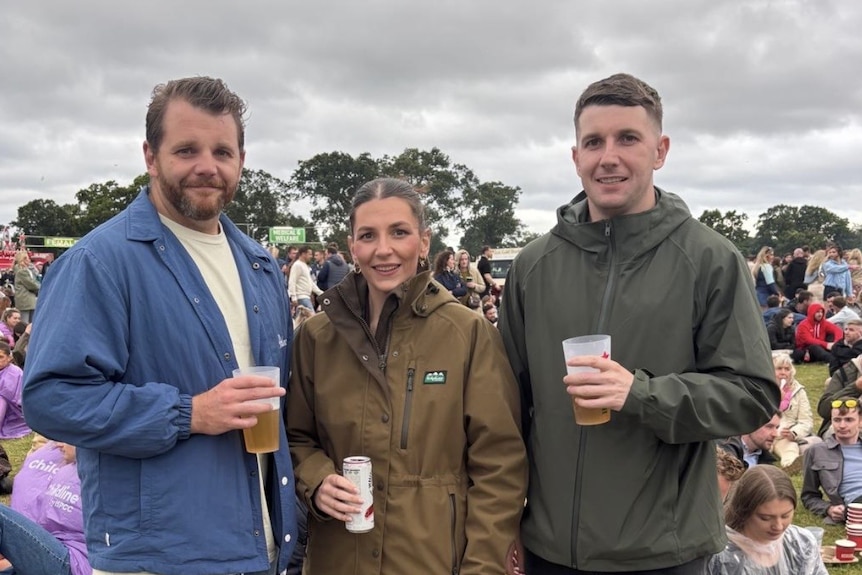 A woman stands between two men, both taller. They are holding drinks and wearing warm jackets.