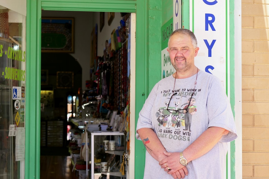 A man in a grey shirt stands in front of a shop doorway.
