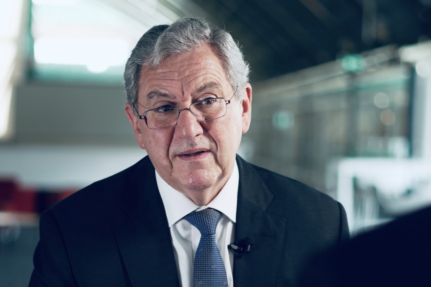 An older man with grey hair and glasses wearing a suit and tie talking in an office.