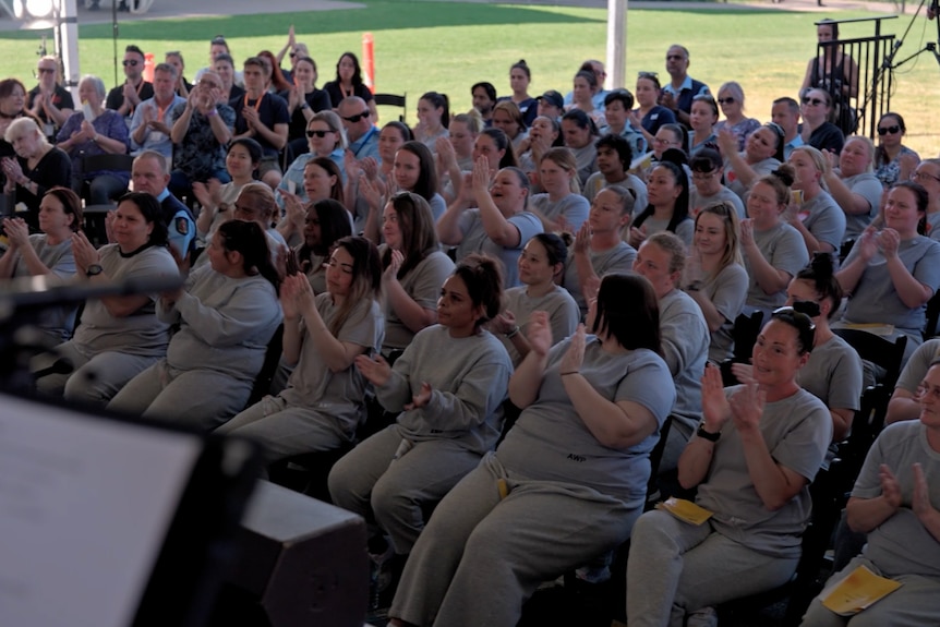 A crowd of about 100 people comprising inmates in gray uniforms applauding, guests and guards, watching a performance.