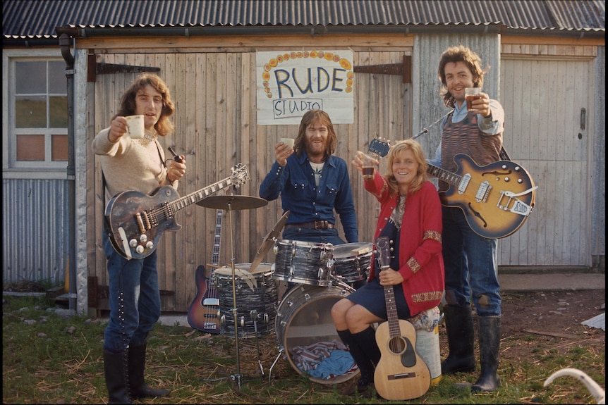 Paul McCartney and Wings members with instruments outside a rustic shed with a handmade "Rude Studio" sign.
