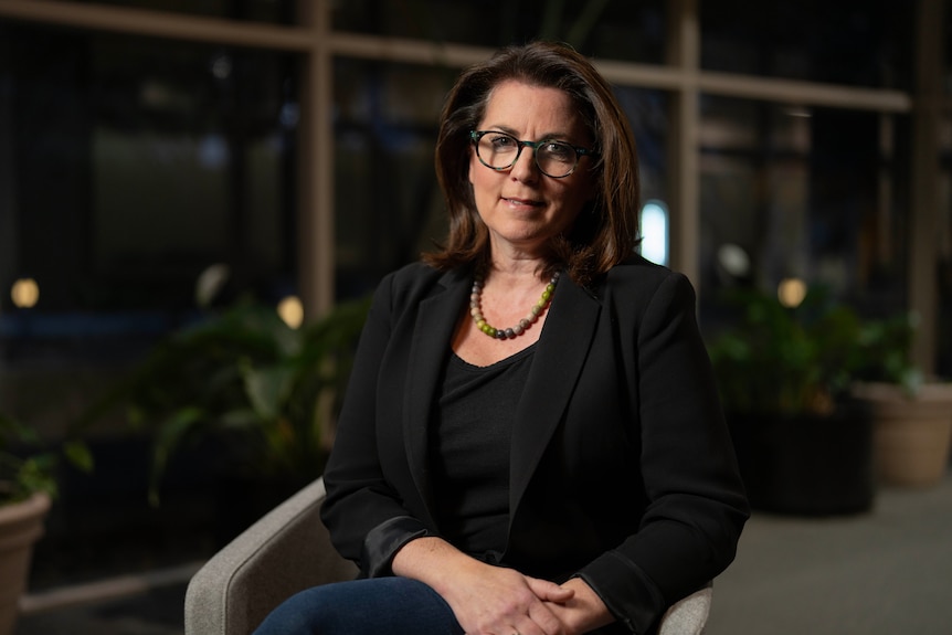 A lady with dark hair, blazer and glasses, sitting in a dark room, smiling at the camera.