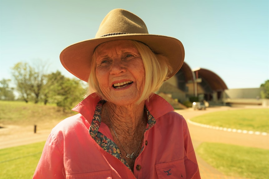 An older, white-haired woman in a broad-brimmed hat stands in a country area.
