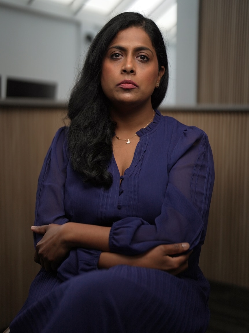 A woman sits indoors looking directly at the camera, she has a serious expression.