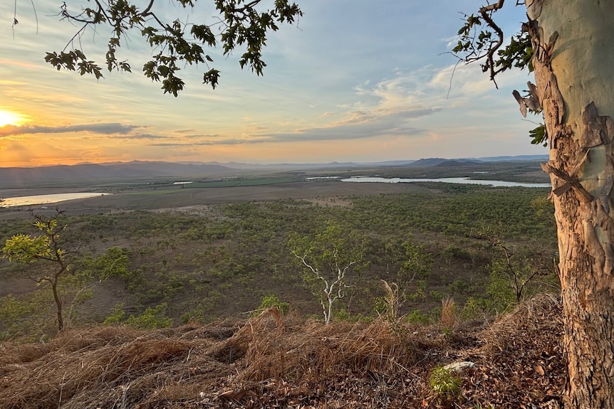 A wide rural landscape stretches to the horizon at sunset, with trees and waterways visible below a hilltop.