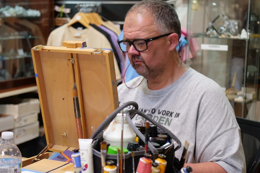 A man in a grey shirt and black-framed glasses looks at a small easel. 