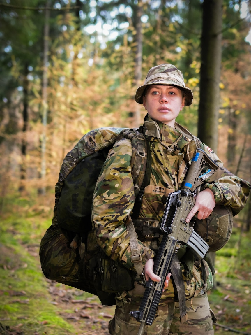 A young woman in khaki camouflage, holding a rifle in the woods.