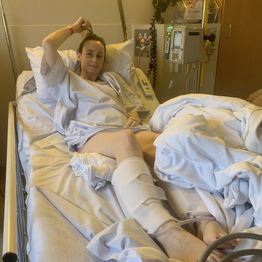 A woman lies in a hospital bed. She is wearing a gown, and looks at the camera, surrounded by some medical equipment.