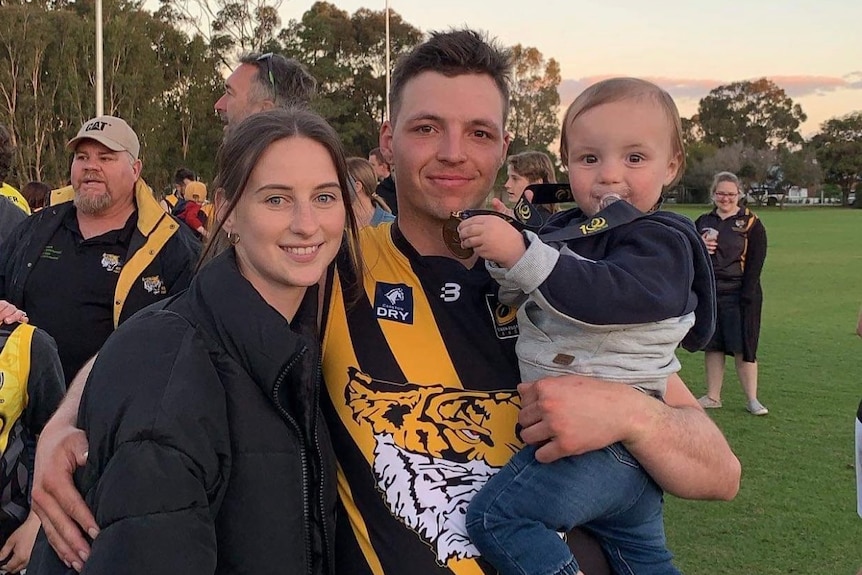 Man in footy jumper with wife and child at a green oval