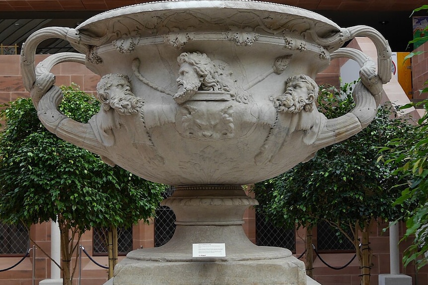 An enormous grey marble urn with handles and decorate faces around the edge.