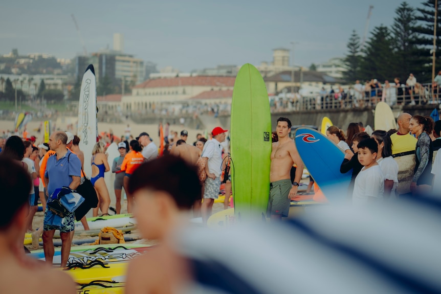 A large crowd of people with paddle boards and surf boards stand on the sand. 